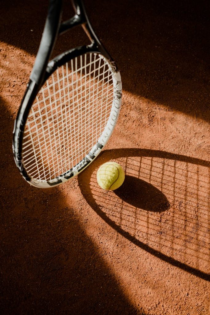 Close-up view of a tennis racket casting a shadow on a clay court with a tennis ball.